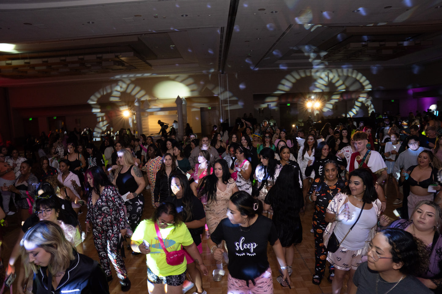 Large crowd dancing and socializing in a dimly lit ballroom with colorful party lights and DJ setup.